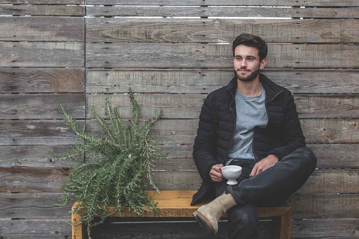 Hombre joven con barba, influencer, bebiendo té, en un banco junto a una bonita planta posando para las redes sociales.